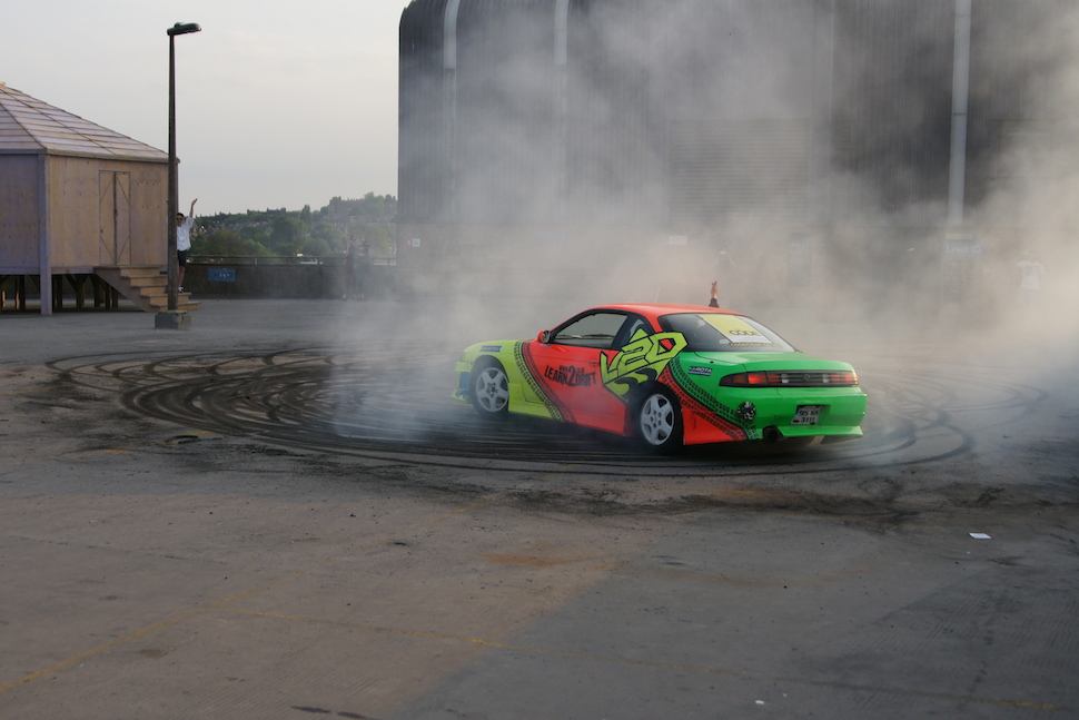 Still from performance of car drawing, drift car in motion making tyre tracks with smoke rising.