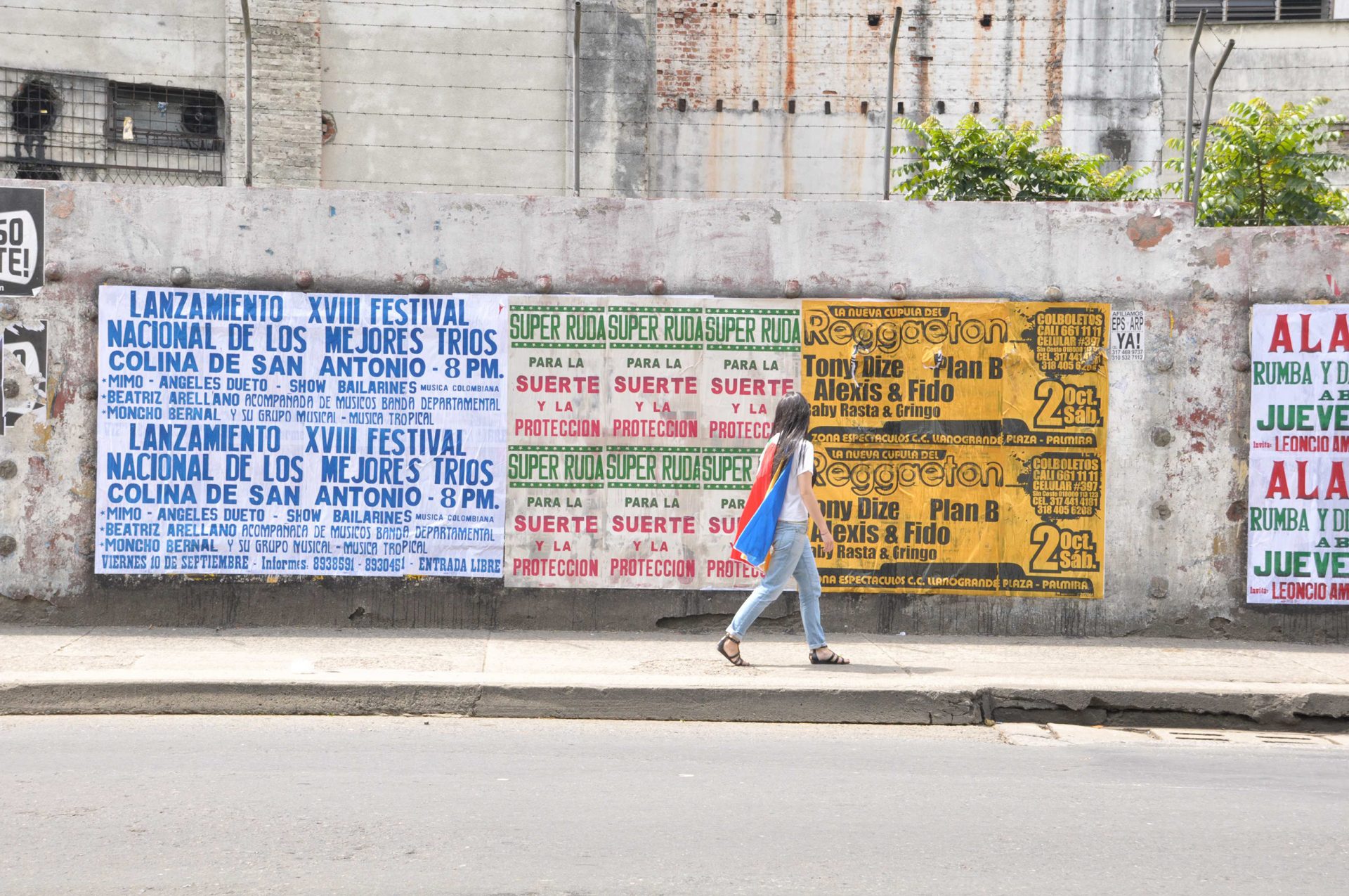 Super Ruda, the alter ego of the artist passes a set of Super Ruda posters pasted on to a wall in cali Colombia. The character is wearing a cape made from a sun umbrella and a fake dark long haired wig. This image is part of a slide show of images from the performance.