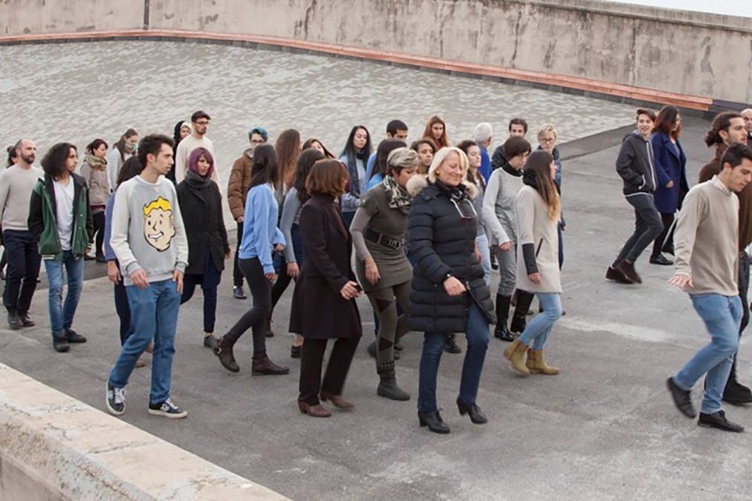 A group of people moving together through a concrete space making the sound of rain with their footsteps.